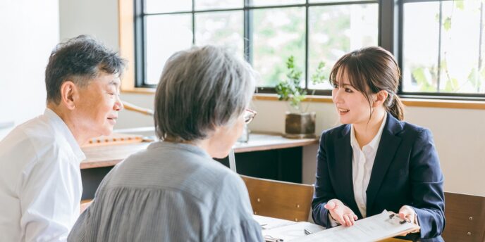 スーツ姿の女性スタッフが高齢夫婦へ住宅相談をしているテーブルシーン