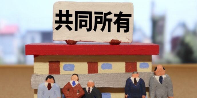 「共同所有」と書かれたプレートが載った建物模型の前に、スーツ姿の人形が複数並んでいる