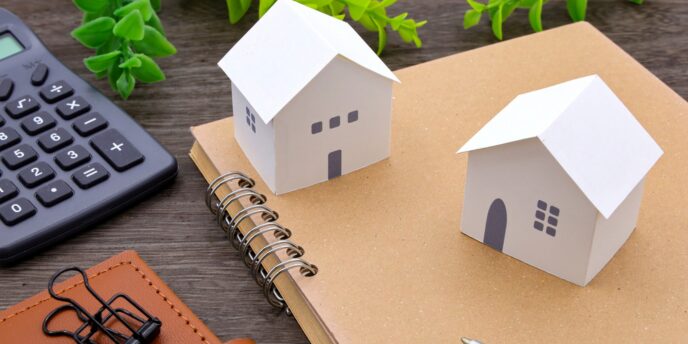 ノートに置かれた紙製の家模型2棟と電卓で住宅資金を検討するイメージ