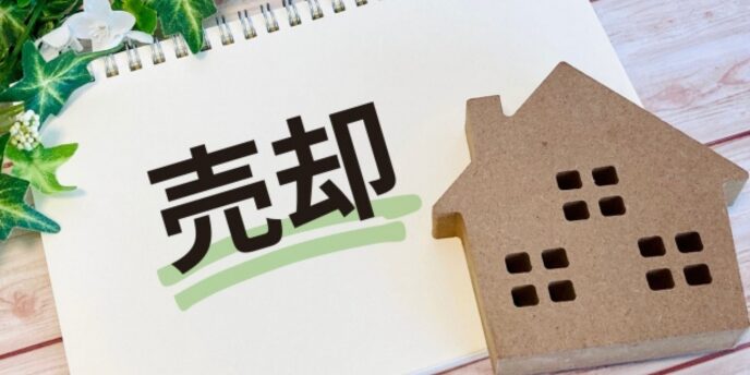 売却」と書かれたノートと家の模型、観葉植物が添えられた不動産売却のイメージ
