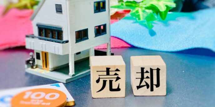 家の模型の前に置かれた「売却」の木のブロックとコインのイメージ